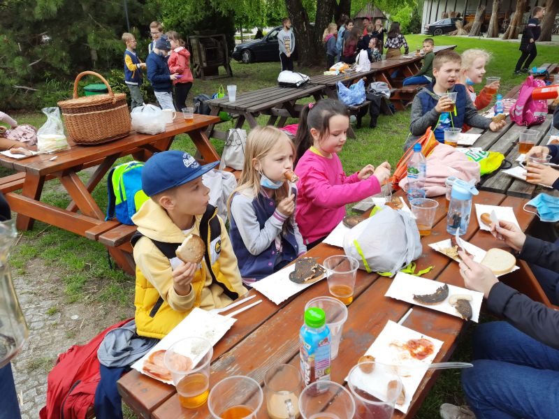 Uczniowie siedzą przy drewnianym stole i jedzą kiełbaski oraz pieczony chleb