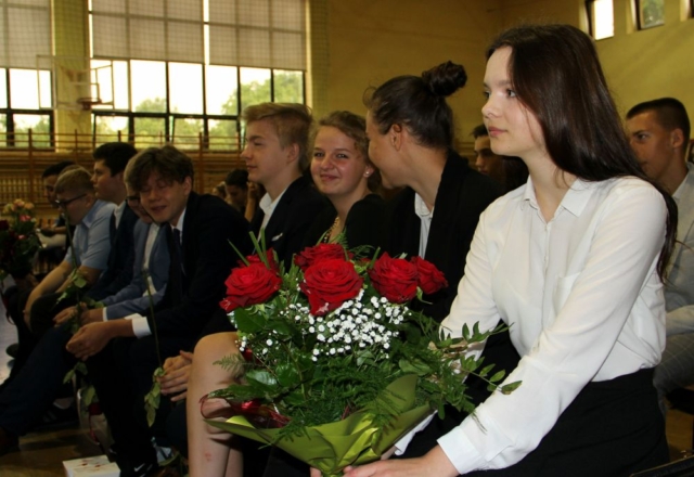 Grupa uczniów siedzi na ławeczkach, trzymają kwiaty