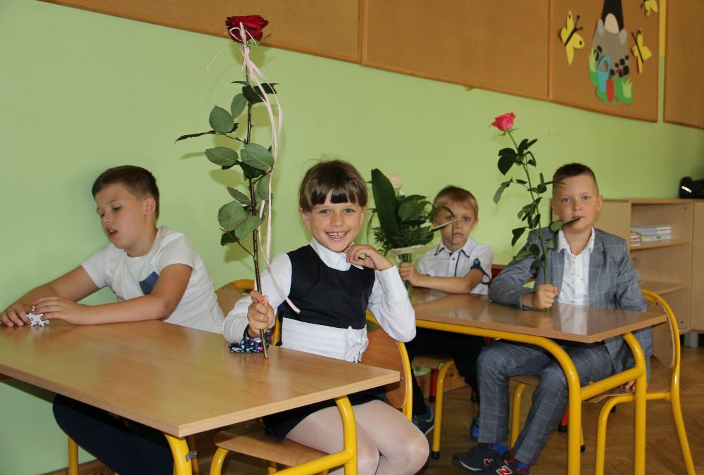 Uczniowie siedzą w klasie w ławeczkach, trzymają kwiaty
