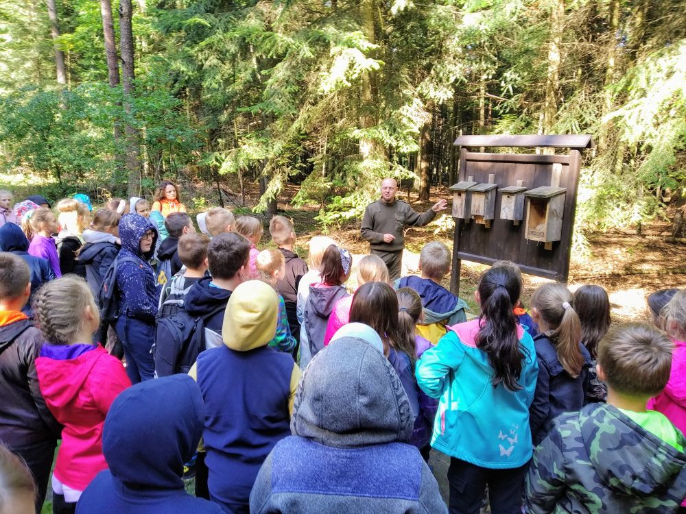 Uczniowie stoją w lesie i słuchają opowieści leśniczego odnośnie budek dla ptaków