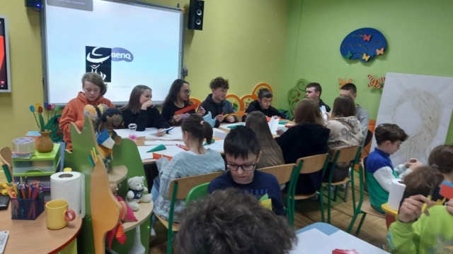 Grupa uczniów siedzi przy stole. Uczniowie wycinają, kleją, tworzą papierowe kwiaty