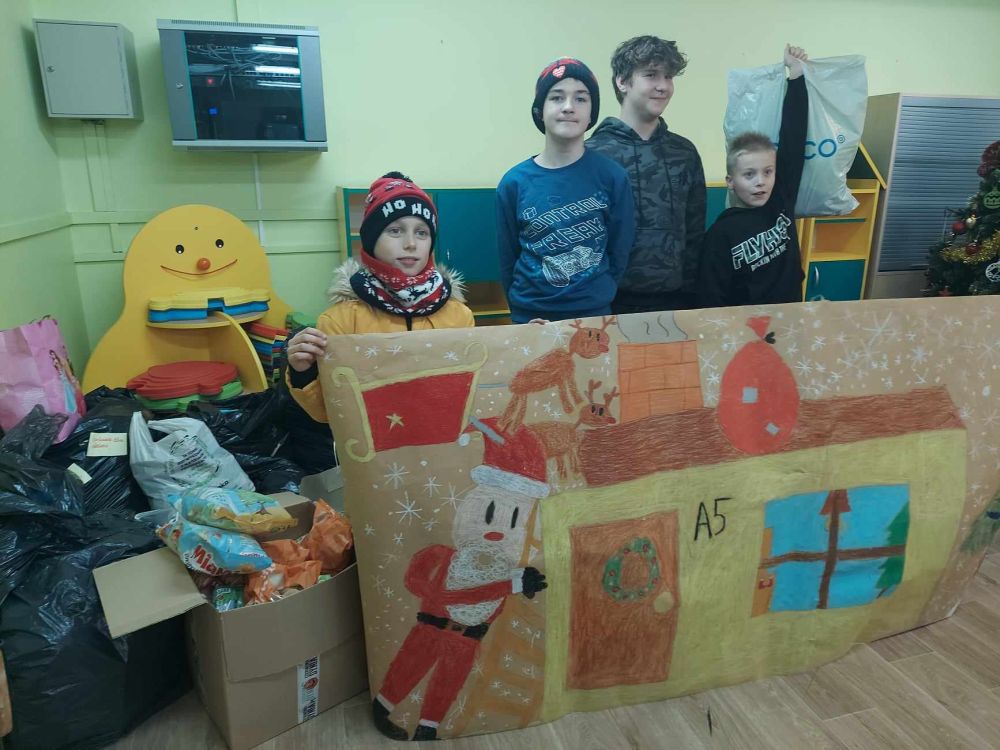 Dzieci stoją przy workach z darami i trzymają plakat z narysowanym domem i Mikołajem